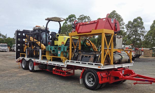 Heavy duty equipment trailer carrying a road-roller, skid-steer and attachments.